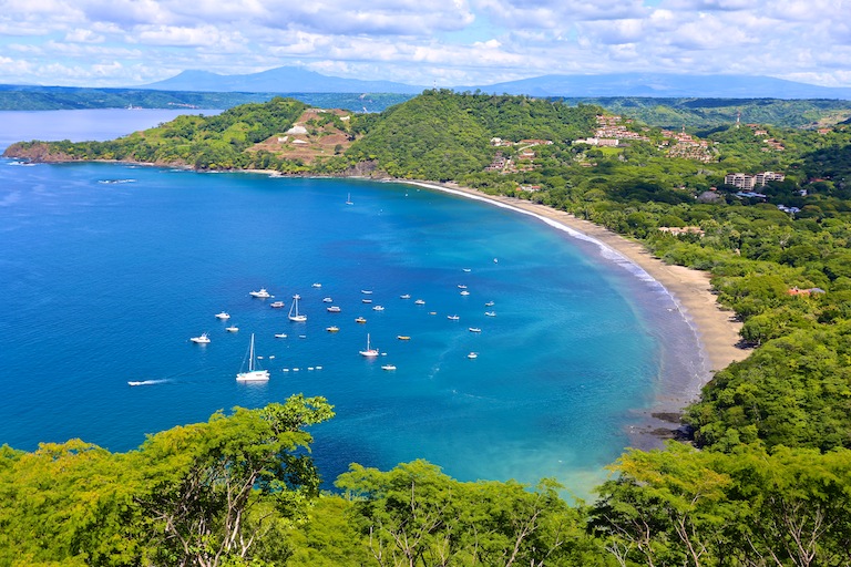 Elevated coastal view of a crescent shaped bay with clear blue water, sailboats at anchor, sandy beach, and dense green hills, highlighting the natural setting and lifestyle appeal of real estate in Costa Rica.