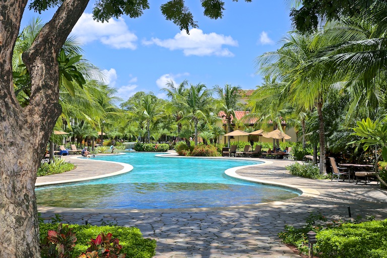 Resort style pool surrounded by palm trees, lush landscaping, lounge seating, and tropical architecture, reflecting relaxed outdoor living within the Costa Rica real estate landscape.