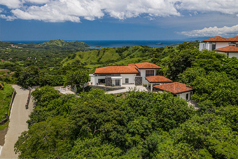 Hilltop home with red tile roofs set above lush green valleys, with a wide ocean horizon and coastal headlands in the distance, capturing elevated view living in the Costa Rica real estate market.