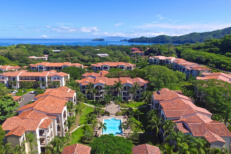 Aerial view of a master planned residential community with tiled roof villas, central pool, tropical landscaping, and ocean views in the distance, highlighting coastal living within the Costa Rica real estate market.